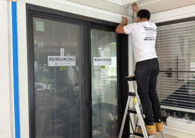 A worker in a white shirt installs a window frame while balanced on a ladder, with 'AG Millworks' signage on the nearby door.