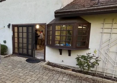 A cozy home entrance featuring a brown bay window, a glass door, and a paved pathway, with workers visible inside.